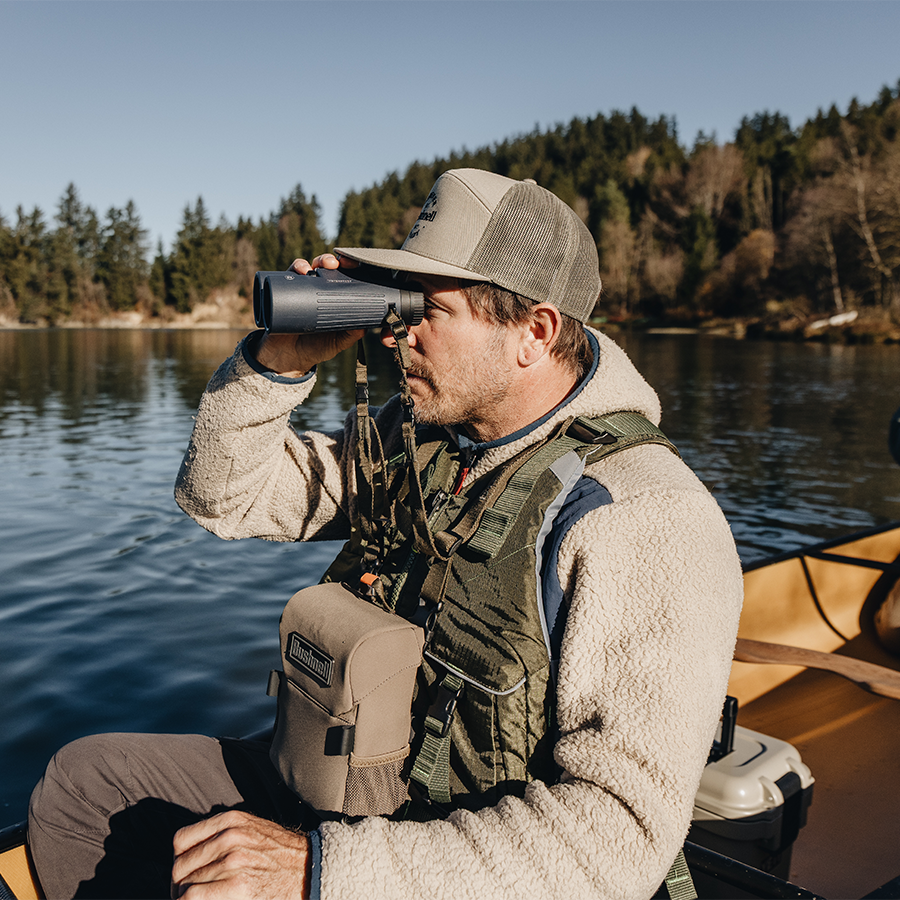 Man in outdoor gear using binoculars by a lake with trees in the background