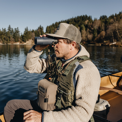 Man in outdoor gear using binoculars by a lake with trees in the background