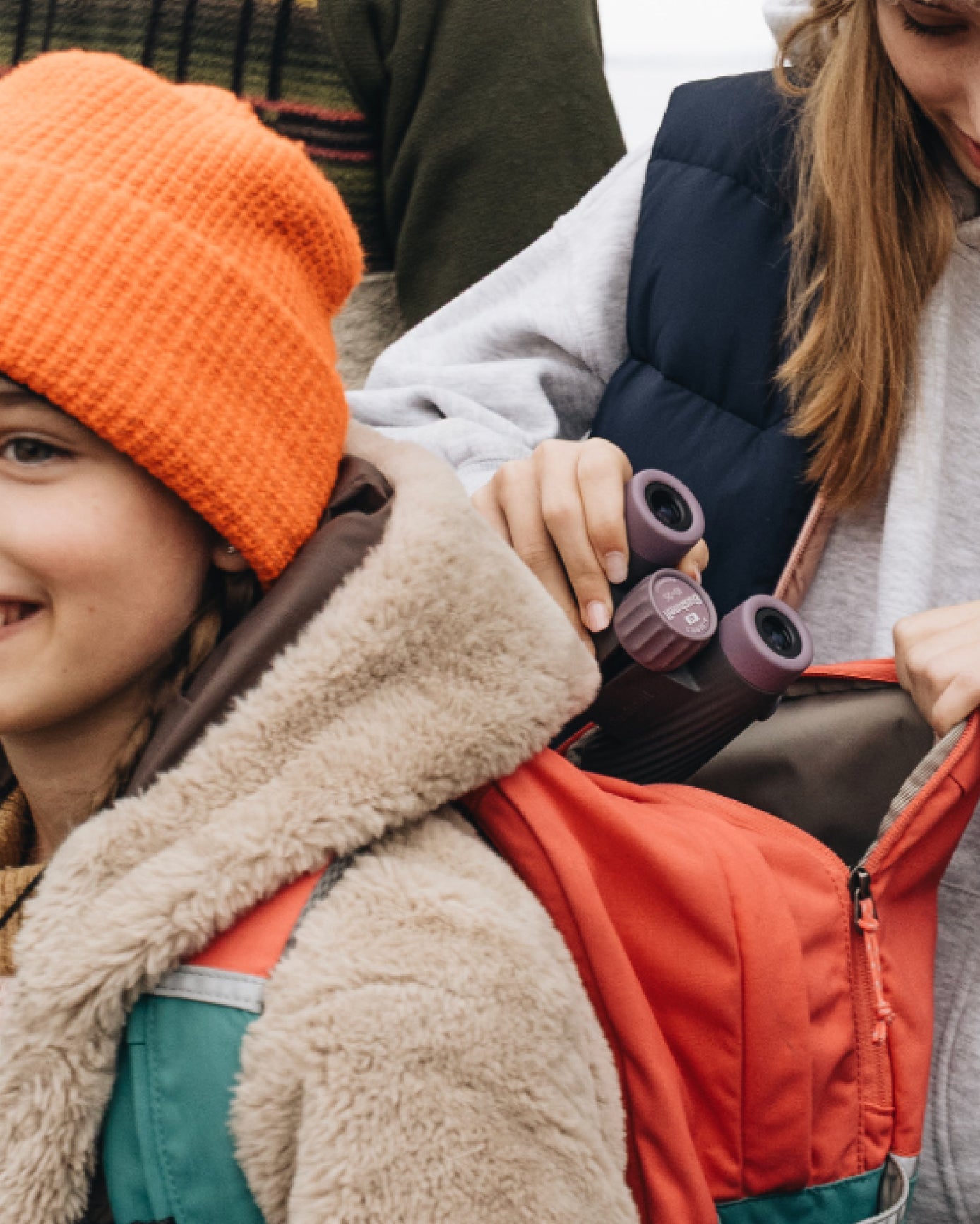 Child wearing an orange beanie and coat, sitting on a person's shoulders, with binoculars held by the person.