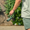 Person holding a green Bushnell A3 monocular by its wrist strap, outdoors with greenery in the background