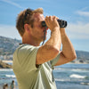 Side view of man using Bushnell A3 binoculars by the ocean with a scenic background