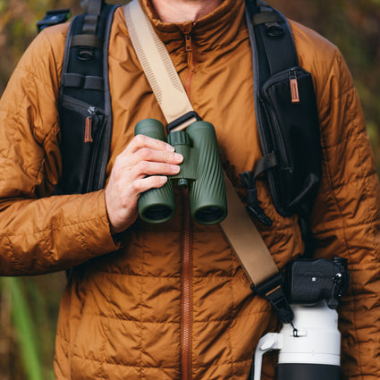 Person in orange jacket holding green Bushnell A5 binoculars with backpack and camera on nature background