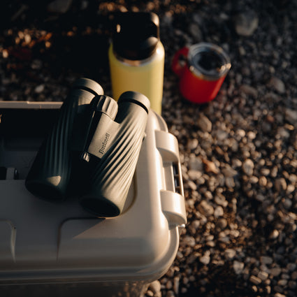 Bushnell A5 binoculars on a cooler with a yellow thermos and red cup in the background on a pebbled riverbank.