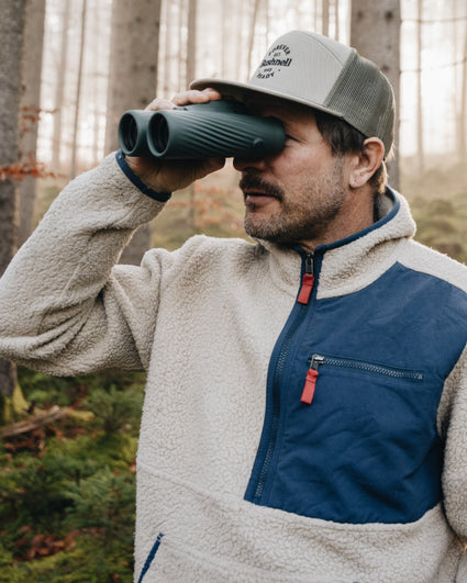 Man in a forest using binoculars, wearing a cap and a two-tone jacket.