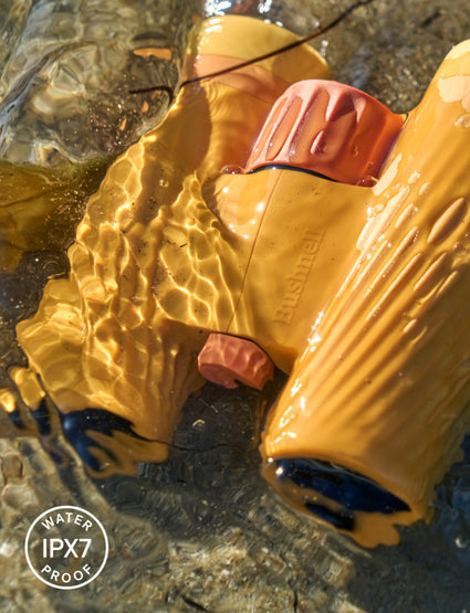 Yellow Bushnell binoculars partially submerged in water with 'Water IPX7 Proof' label.