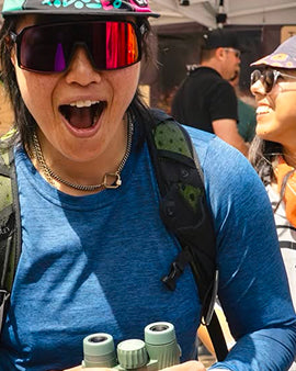 Person wearing a helmet and sunglasses with binoculars, surrounded by people in an outdoor setting