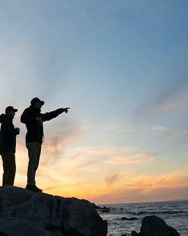 Two people standing on rocks by the ocean at sunset, silhouetted against the sky.