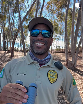 Person wearing a uniform with a badge, holding binoculars, in a forested area.